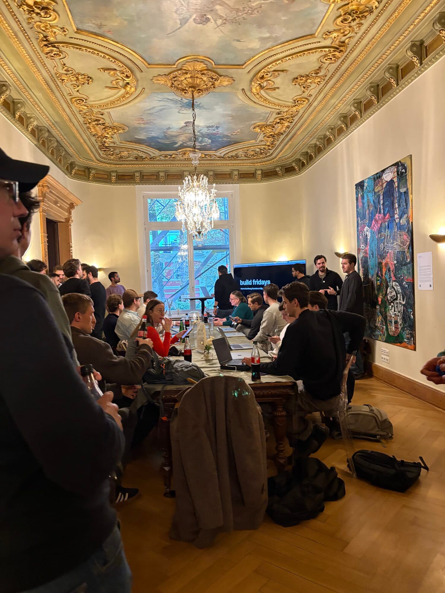Members gathered around tables in a grand room with a painted ceiling and chandelier at an AI BEAVERS event