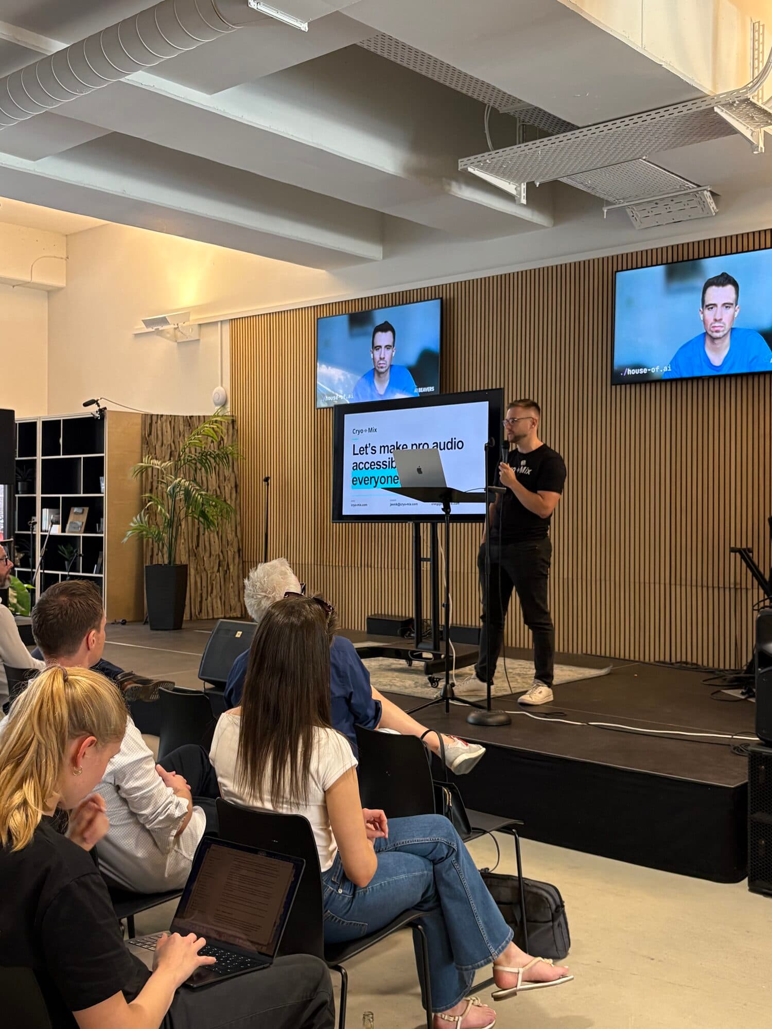 A host on stage beside two screens while members listen from low sofas at an AI BEAVERS event