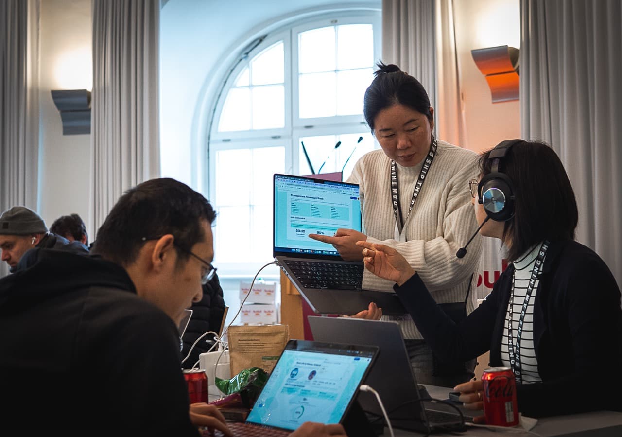 A builder standing at her team's laptops by a bright window at the AI BEAVERS January 2026 hackathon in Hamburg