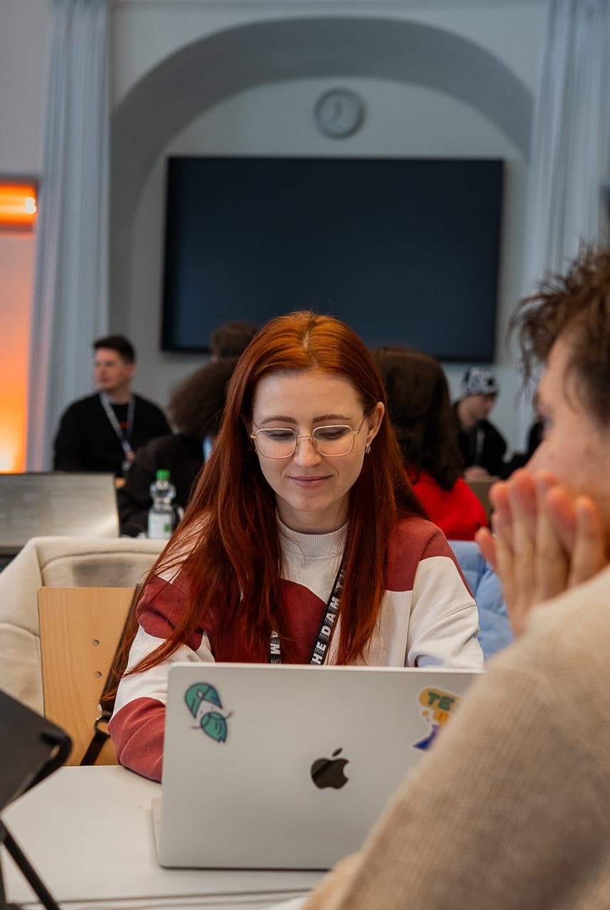 A builder with red hair smiling at her team's table at the AI BEAVERS January 2026 hackathon in Hamburg