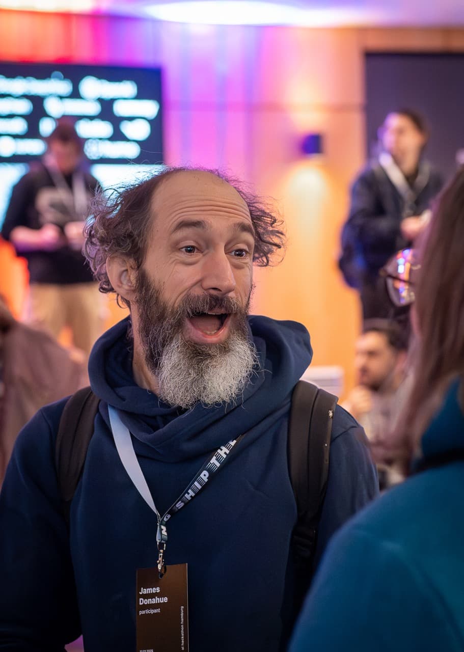 A grey-bearded attendee grinning in the crowd at the AI BEAVERS January 2026 hackathon in Hamburg